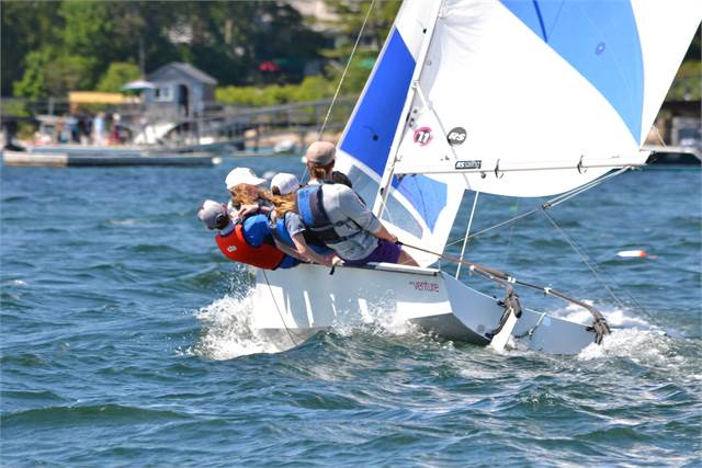 Three people wearing life jackets and caps sail in a small white sailboat with blue-and-white sails, leaning out over the water as the boat tilts, with a shoreline and docks in the background.