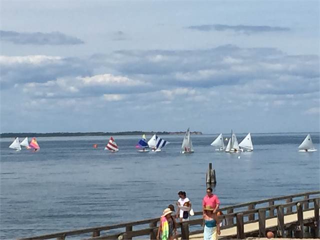 Several small sailboats with colorful sails are on a calm body of water under a partly cloudy sky, while people stand and walk on a wooden pier in the foreground.