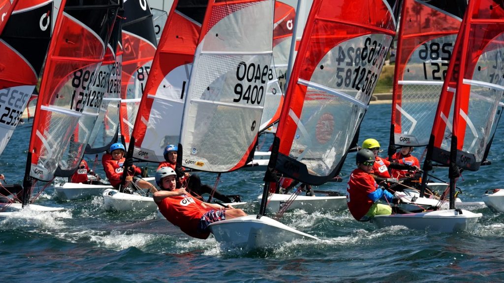 A group of young sailors wearing helmets and red shirts compete in a sailing race, navigating small boats with red and white sails on a sunny day. The water is choppy, and the background shows land and a fence.
