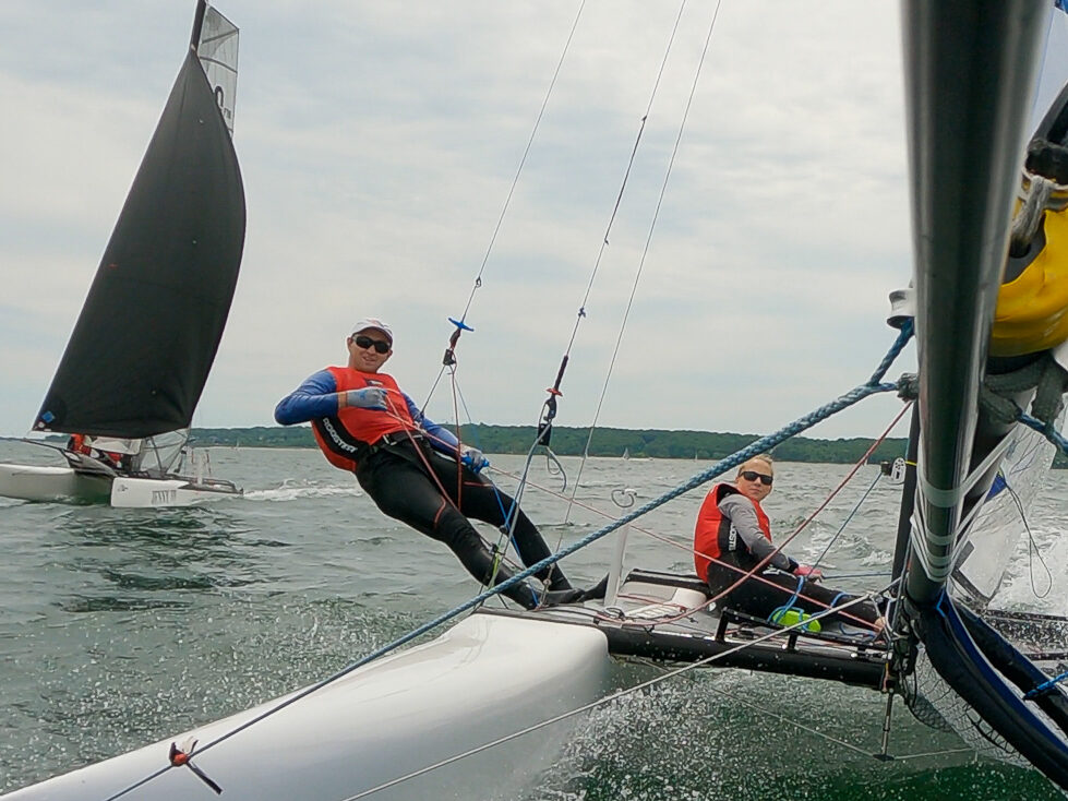 Two people in life jackets sail a small boat on the water; one leans out over the edge while the other steers. Another sailboat with a black sail is visible in the background. Trees line the distant shore under a cloudy sky.
