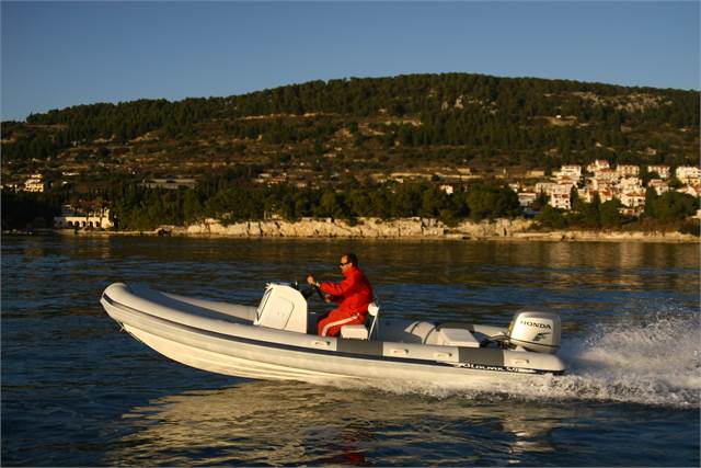 A person in a red outfit drives a white inflatable motorboat with a Honda engine across calm water, with a forested hillside and buildings in the background under a clear sky.
