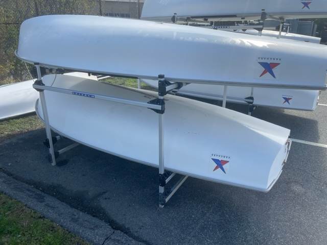 Two white sailboats are stored upside down on a metal rack in an outdoor area. Both boats display a blue and red 'X' logo on the hull. The scene is well-lit with sunlight.