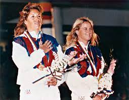Two female athletes in white tracksuits with red and blue details stand side by side, each holding flowers and wearing medals around their necks, smiling during an award ceremony.