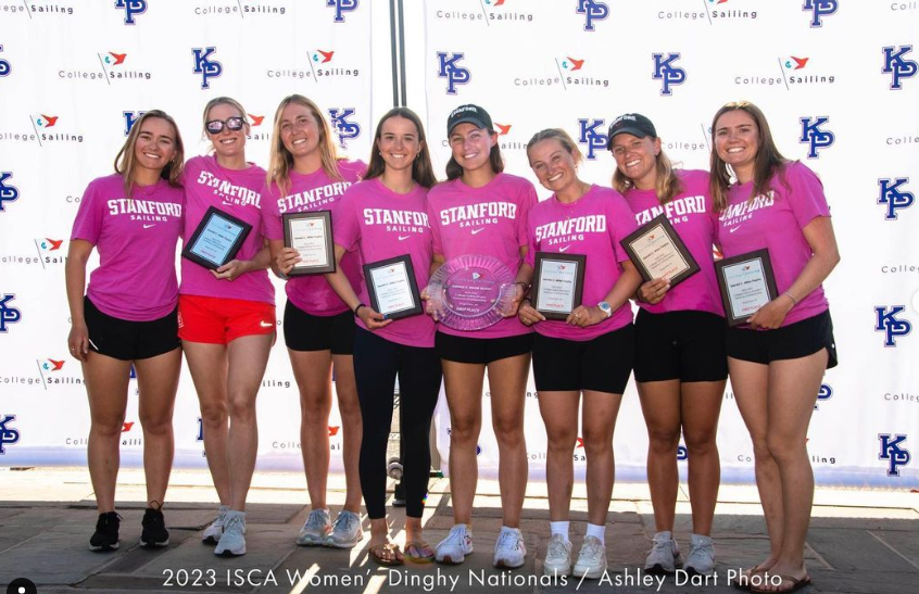 Eight women wearing matching pink 'Stanford Sailing' shirts and black shorts stand smiling, holding plaques and trophies, in front of a backdrop with sailing and college logos. Text at bottom reads '2023 ISCA Women Dinghy Nationals.