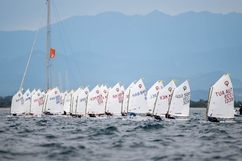 A fleet of small sailboats with white sails, each marked with numbers and letters, race across choppy water under an overcast sky with mountains in the background.