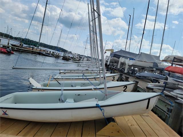 A row of small white sailboats is lined up on a wooden dock next to a marina, with more boats and masts visible on the water under a partly cloudy sky.
