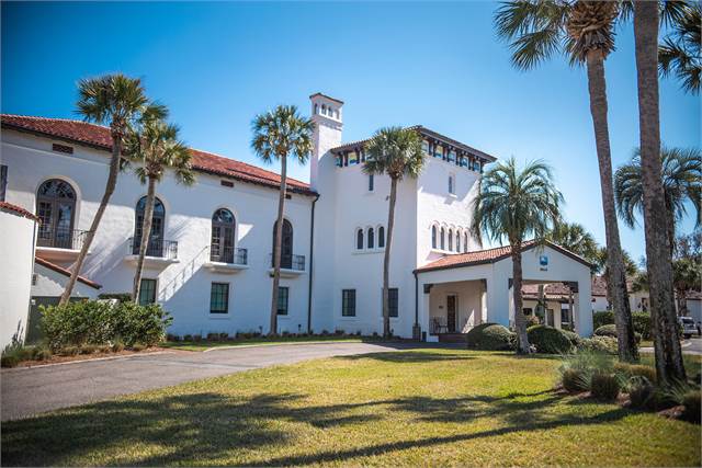A large white stucco building with red-tiled roofs, arched windows, and a central tower, surrounded by palm trees and a manicured lawn on a sunny day.