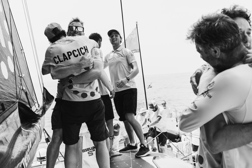 A group of sailors celebrate on a boat, hugging and smiling. One person’s shirt reads 'CLAPCICH.' The scene is energetic and joyful, with the ocean visible in the background. The image is in black and white.
