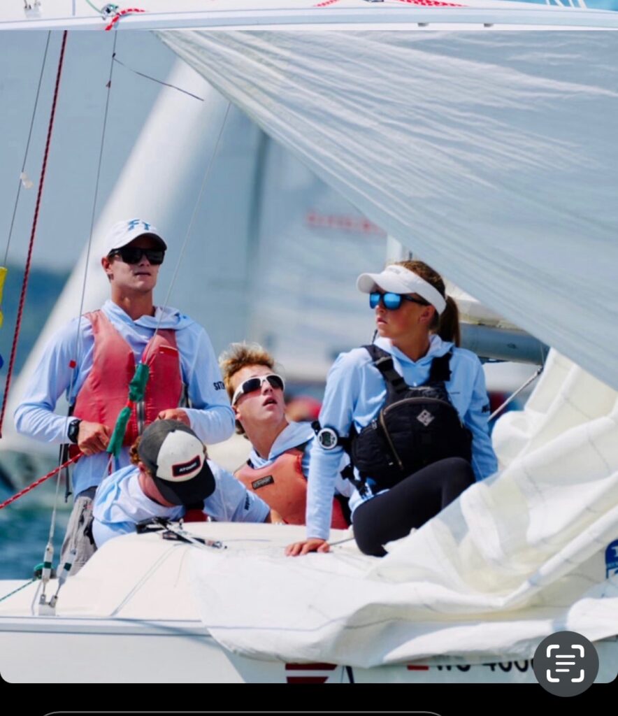 Four people in sailing gear are on a sailboat, focused on adjusting the sails. One person is looking up, another looks forward wearing sunglasses, and two are handling lines, with the sea and other boats in the background.