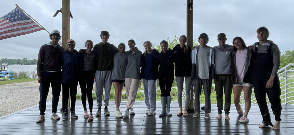 A group of thirteen teens stand side by side under a wooden shelter near a lake, smiling with arms around each other. An American flag and green trees are visible in the background on a cloudy day.