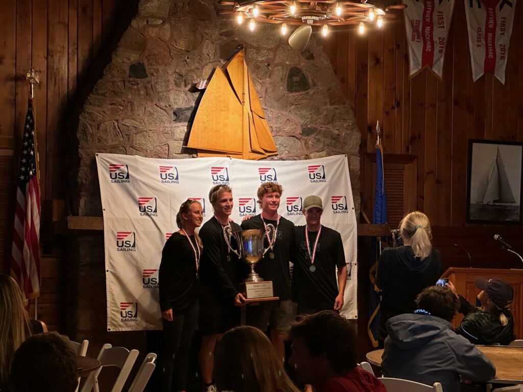 Four people stand together holding a large trophy and wearing medals in front of a USA Sailing backdrop inside a wooden lodge, while others look on and take photos. A sailboat model and flags are displayed in the background.