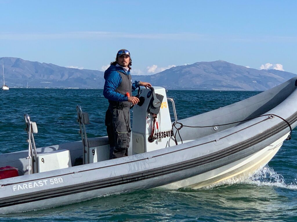 A person stands at the helm of a gray inflatable motorboat on the water, with distant mountains and a sailboat visible in the background under a clear blue sky.