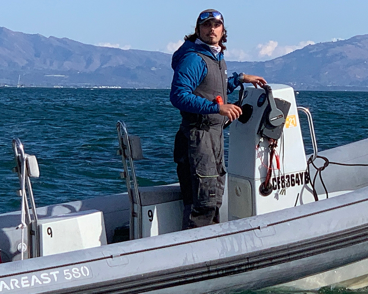 A person wearing a blue jacket, life vest, and sunglasses stands at the controls of a grey inflatable motorboat on open water, with mountains visible in the background.