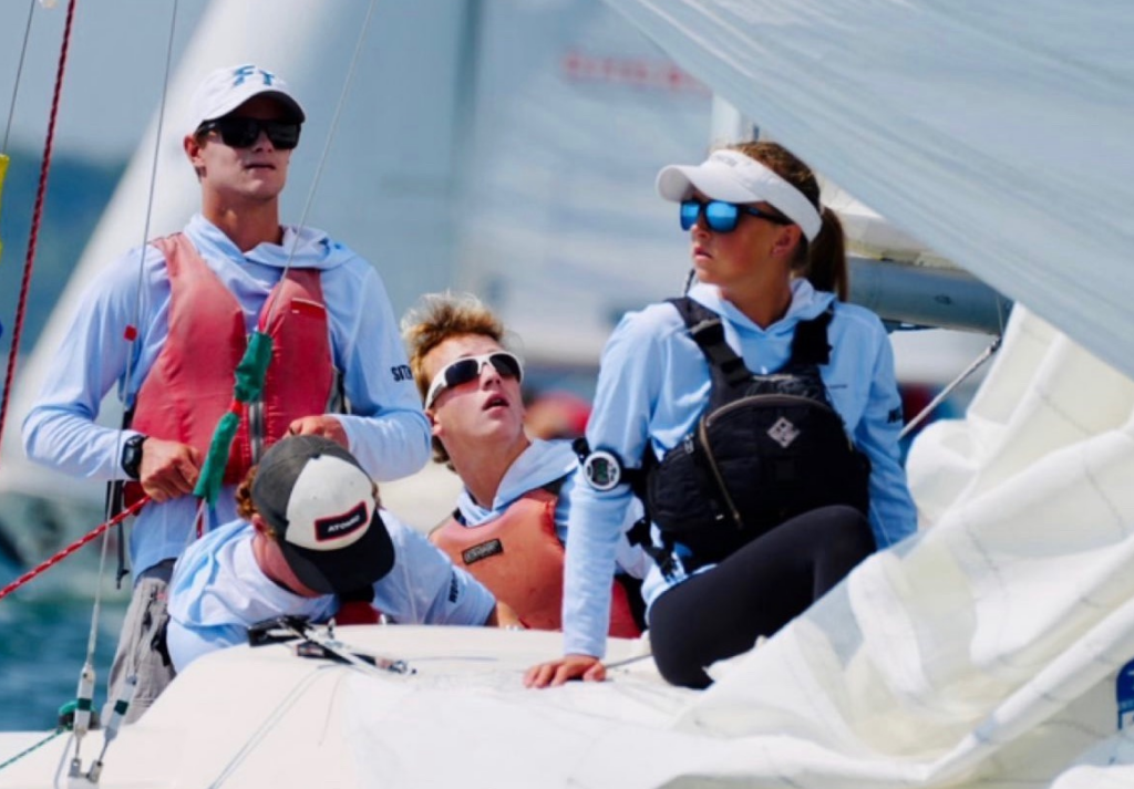 Four young sailors, wearing sunglasses and life vests, are working together on a sailboat. They appear focused, adjusting sails and ropes as they navigate on the water under bright daylight.