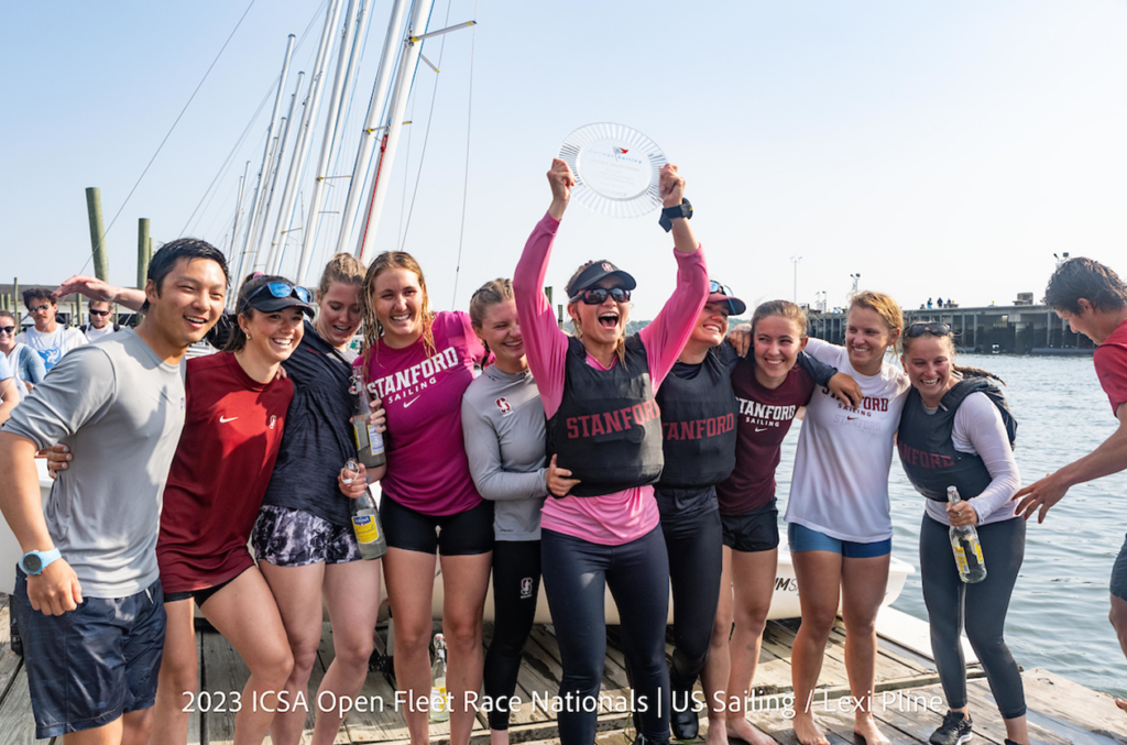 A joyful group of Stanford sailing team members stand together on a dock, smiling and celebrating as one holds up a glass trophy plate at the 2023 ICSA Open Fleet Race Nationals.