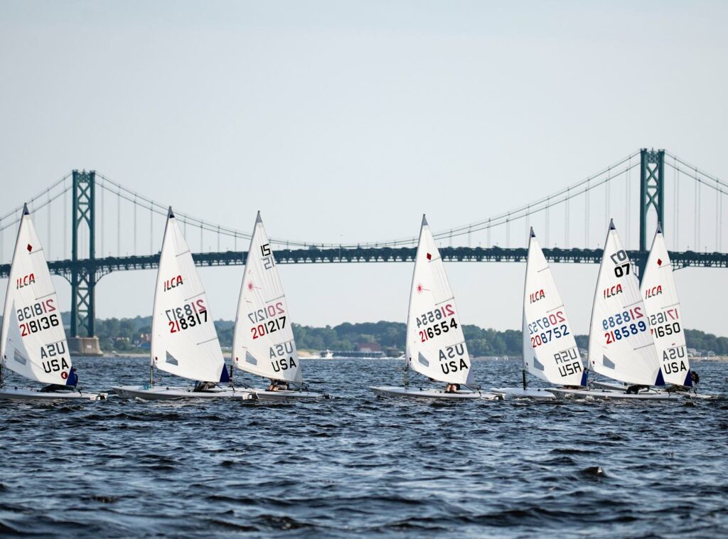 Several small sailboats with white sails and marked with “USA” race on the water, with a large suspension bridge and distant shoreline visible in the background under a clear sky.