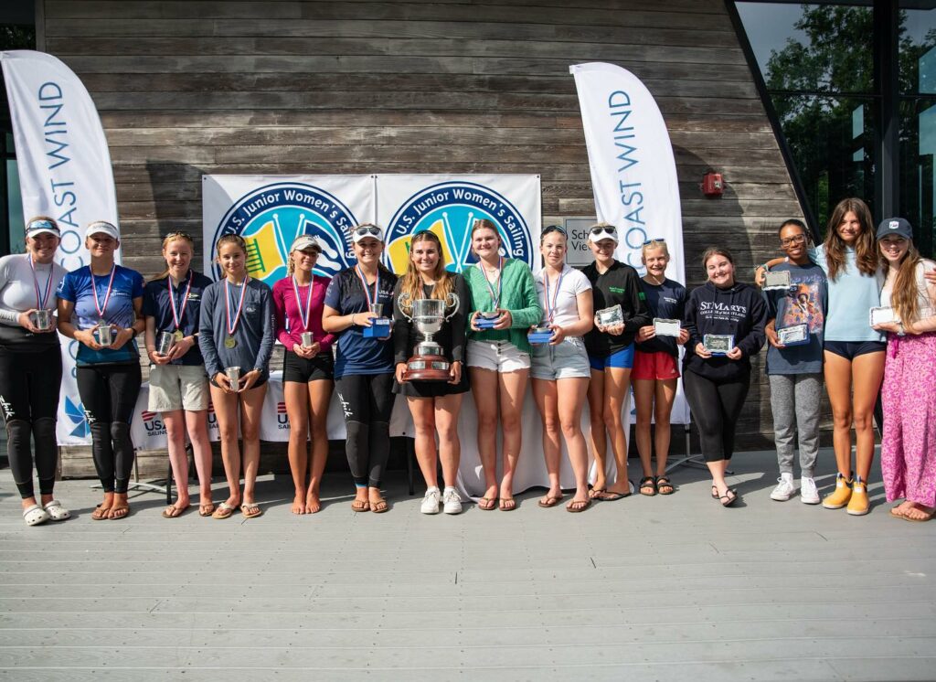A group of young women stand in a row on a wooden deck, holding trophies and medals. Two banners and a sign with “US Junior Women’s” are visible behind them. All are smiling in casual and athletic wear.