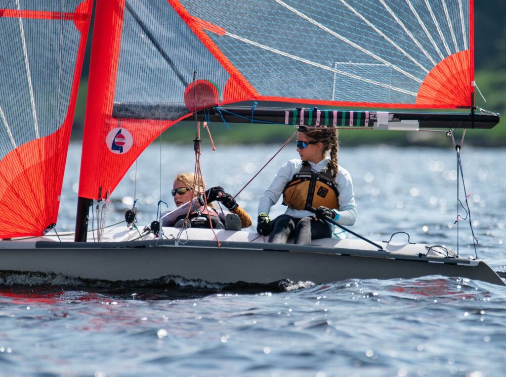Two young sailors wearing life jackets and sunglasses navigate a small sailboat with bright red sails on a calm body of water, with greenery visible in the background.