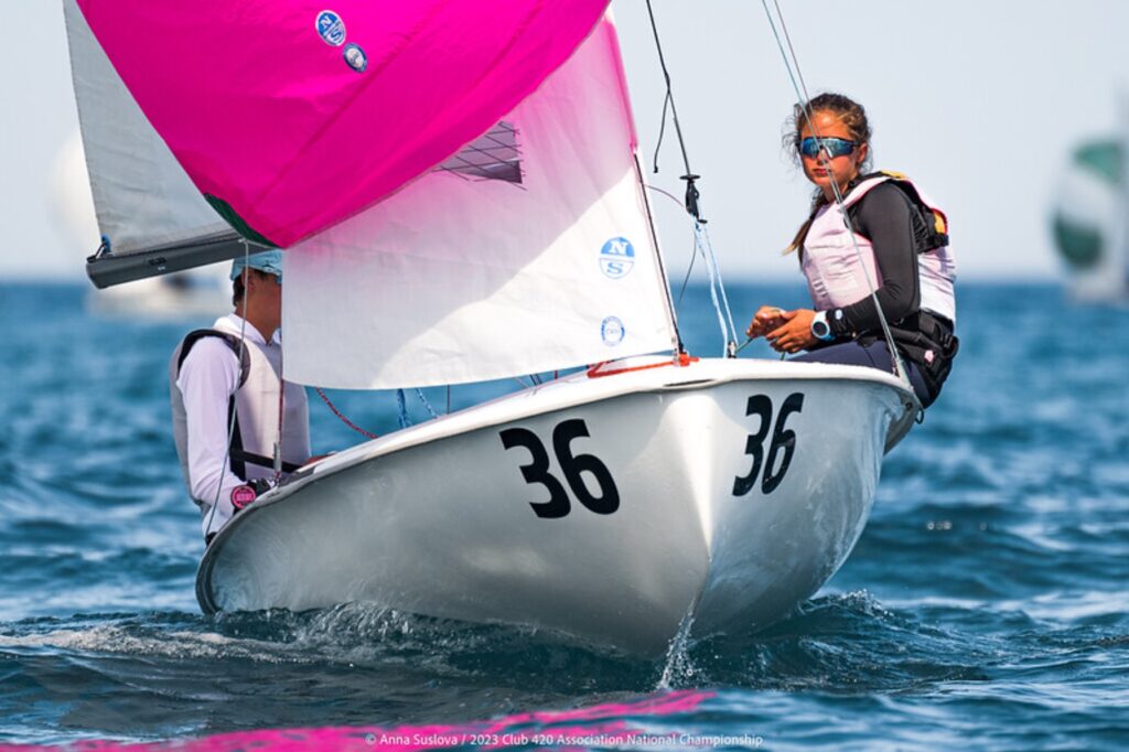 Two young sailors in life jackets race a small sailboat with number 36, featuring a bright pink sail, on blue water during a sunny day. One sailor is sitting on the edge, steering and looking ahead.
