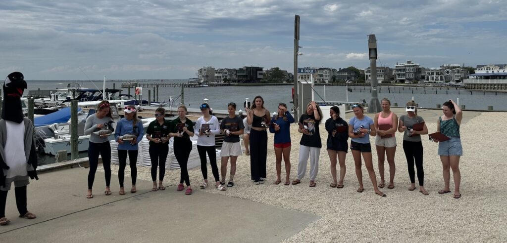 A group of people stands in a line outdoors near a marina, each holding a small trophy. Boats and waterfront houses appear in the background under a cloudy sky. One person on the left wears a penguin costume.