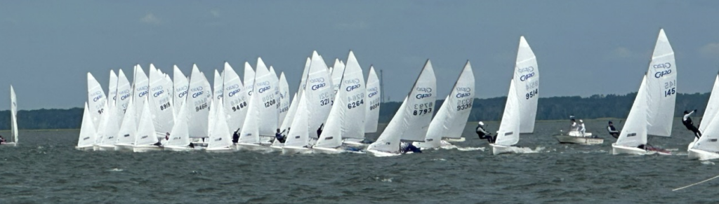 A group of small sailboats with white sails are racing closely together on a choppy body of water under a partly cloudy sky, with a few motorboats in the background.