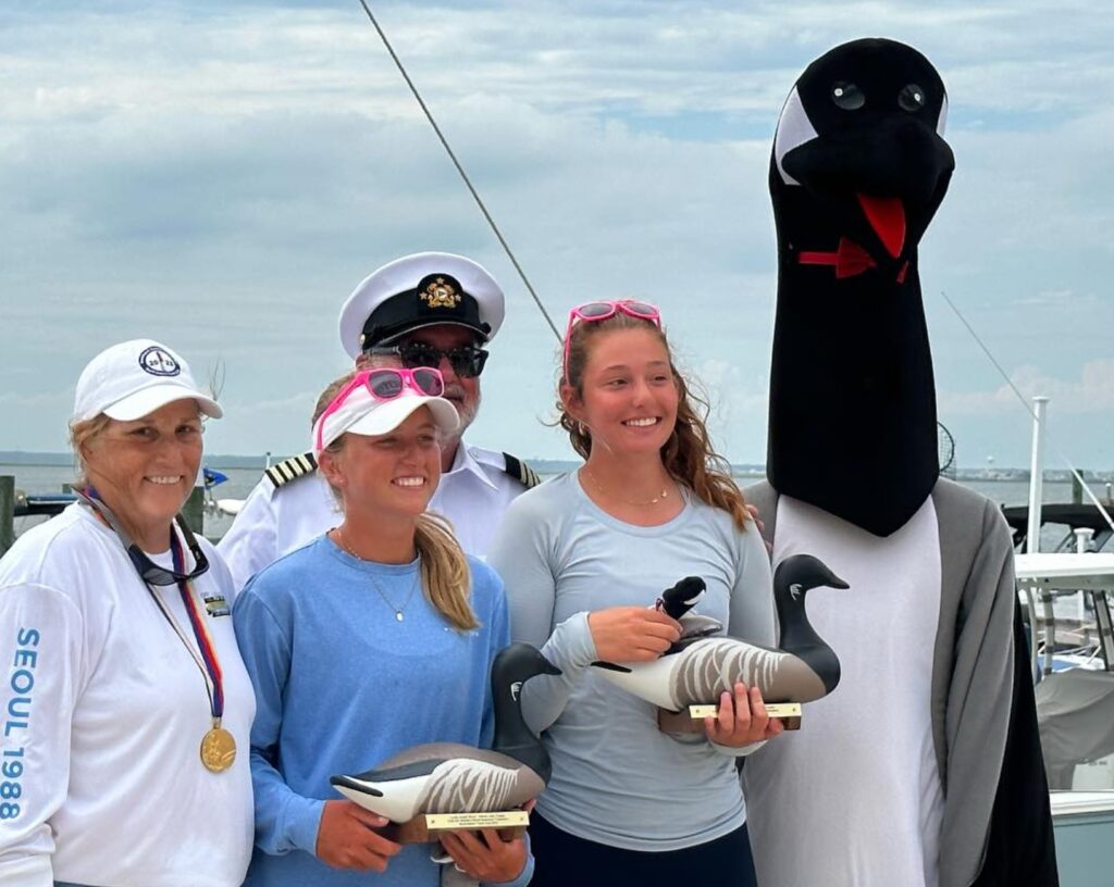 Four people stand on a dock, two holding goose-shaped trophies, with a fifth person dressed in a goose costume. The group smiles and the sky is cloudy, boats are visible in the background.