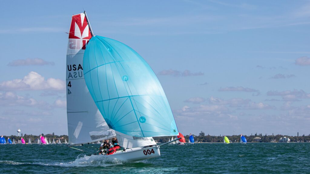 A white sailboat with a large blue spinnaker sail glides across the water. Multiple people are on board, and “USA 4” is visible on the sail. Other sailboats are in the background under a blue sky.