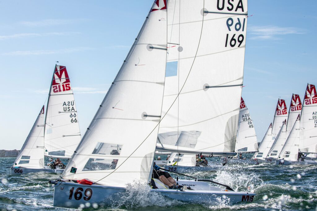 A group of small sailboats with white sails, each marked with numbers and 'USA,' racing on choppy water under a clear blue sky. Sailors are actively navigating through splashing waves.