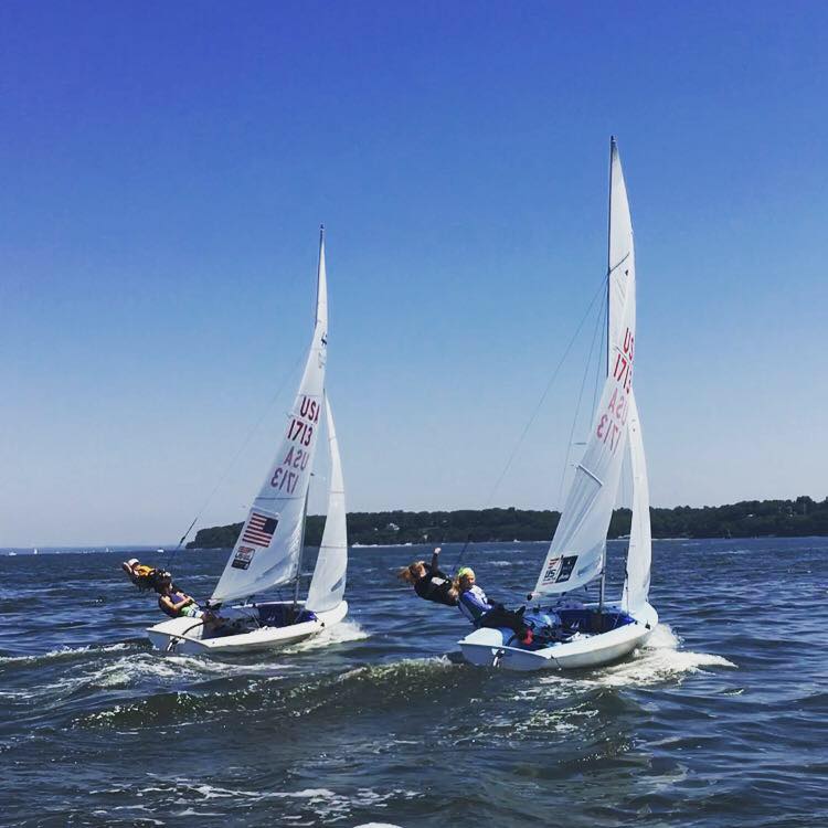 Two sailboats with teams of sailors leaning out over the water race side by side on a sunny day, with a tree-lined shore visible in the background under a clear blue sky.
