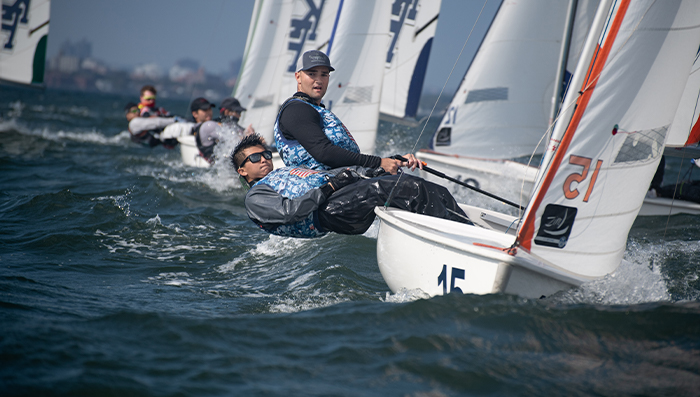 Two sailors in blue vests lean out of a small sailboat, racing on choppy water with several other boats behind them. The city skyline is visible in the background under a clear sky.