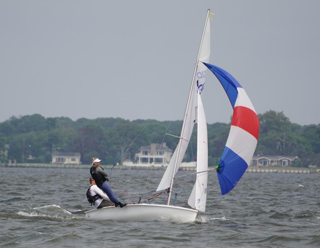 Two people sail a small boat with a colorful red, white, and blue spinnaker on a body of water, leaning out to balance the boat. Houses and trees are visible along the distant shore under a cloudy sky.