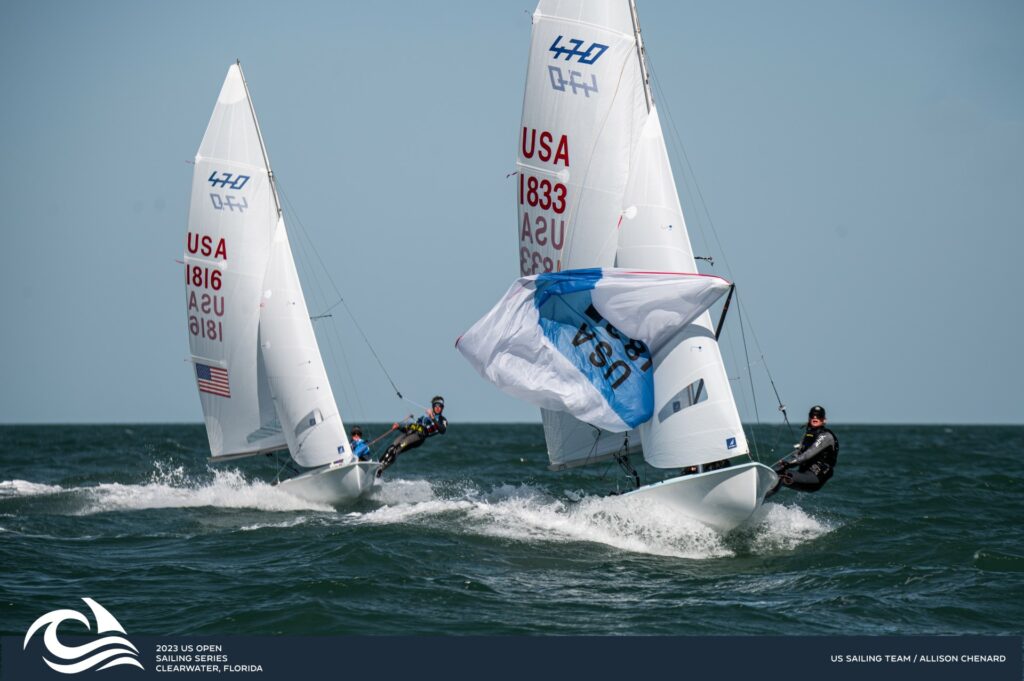 Two sailboats with "USA" sails race side by side on the ocean during sunny weather, with sailors leaning out. Event branding and wave graphics appear in the lower left corner.