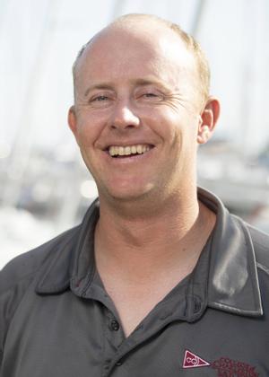 A smiling man with short light hair wearing a gray collared shirt with a red and white emblem, standing outdoors with blurred boats and masts in the background.