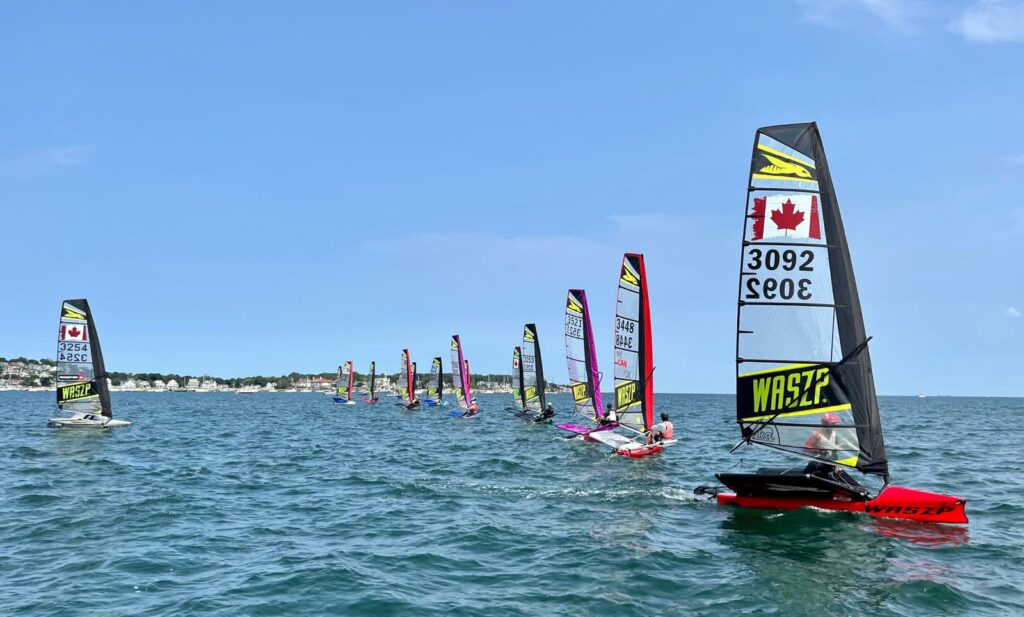 A group of small sailboats with colorful sails, many displaying Canadian flags, race on blue water near a distant shoreline under a clear sky.