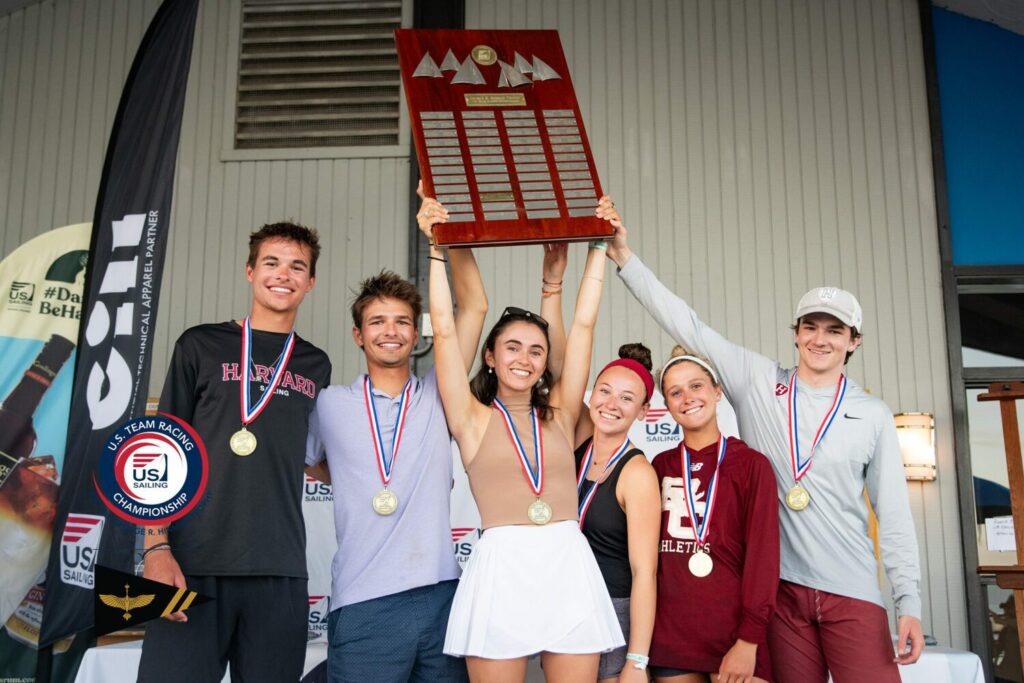 Six young sailors wearing medals stand together, smiling. The woman in the center lifts a large trophy plaque overhead. They pose in front of nautical banners, celebrating their victory at a sailing championship.