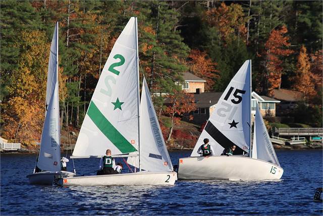 Two small sailboats, one with a green '2' and one with a black '15' on their sails, race on a lake bordered by trees with autumn foliage and houses in the background.
