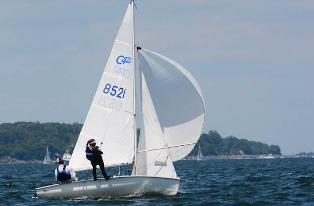 Two people sail a small white sailboat with the number 8521 on the sail, gliding on a body of water with green trees and a few other boats in the background under a blue sky.