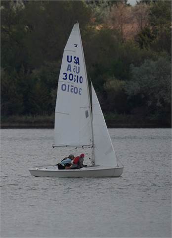 Two people are sailing a small white sailboat with the sail marked 'USA 30510' on a calm body of water, with trees and greenery visible in the background.