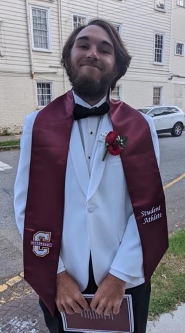 A young man in a white tuxedo and maroon sash labeled 'Student Athlete' stands outside, smiling, holding a diploma. He has a boutonniere on his jacket and an old white building and car are in the background.