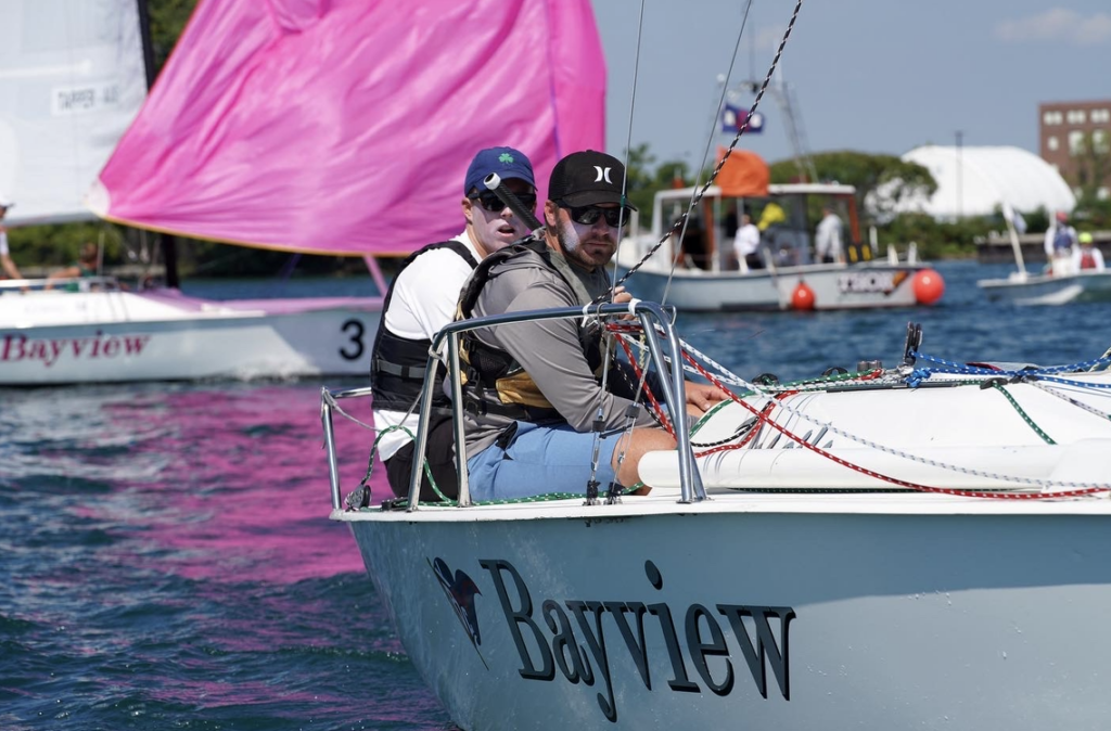 Two people sailing a white boat labeled 'Bayview' on a sunny day, with a pink sailboat and another boat in the background on blue water. The sailors wear hats and sunglasses.