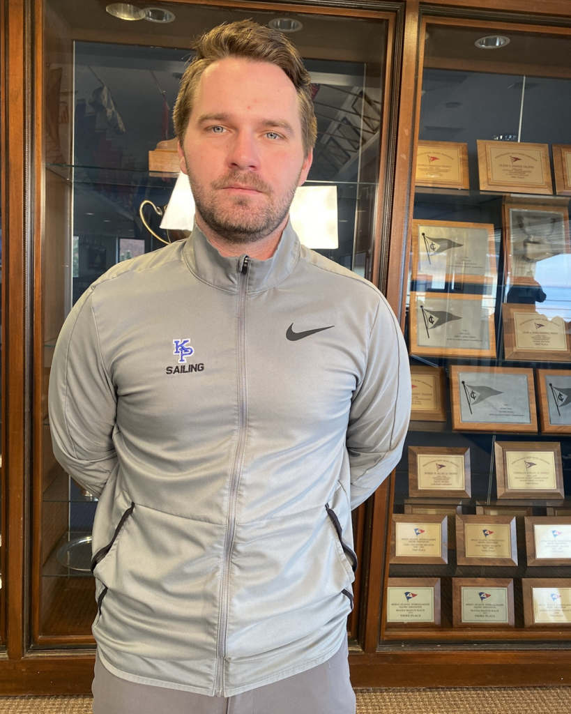 A man in a gray KP Sailing Nike jacket stands indoors in front of a glass display case filled with award plaques. He has short brown hair and a neutral expression, with his hands behind his back.