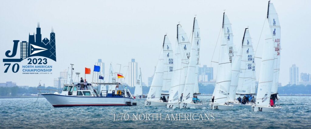 A group of sailboats with white sails racing on a lake, accompanied by a committee boat with colorful flags; city buildings and the 'J/70 2023 North American Championship' logo are visible in the background.