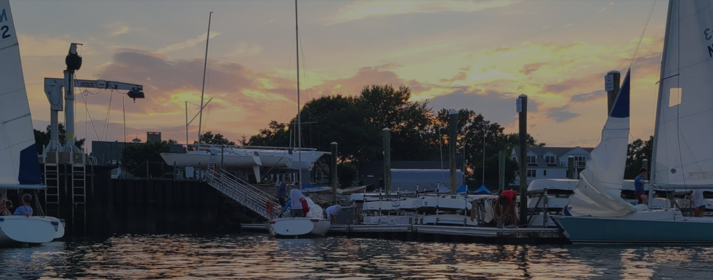 Sailboats docked at a marina during sunset, with people standing on the dock. The sky is partly cloudy with warm orange and yellow hues, and trees and houses are visible in the background.