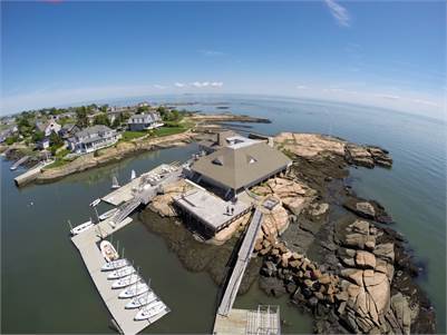 Aerial view of a coastal marina with a central building on rocky shore, boats docked at a pier, and surrounding houses overlooking calm ocean waters under a blue sky.