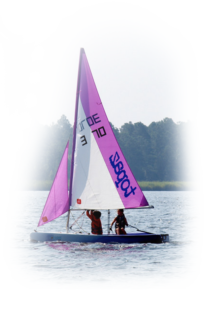 Two people sail a small boat with purple and white sails on a calm lake, with trees and greenery visible in the background.