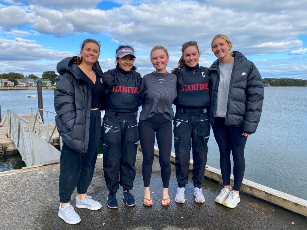 Five young women stand side by side on a dock by the water, dressed in warm jackets and athletic gear, smiling at the camera with a cloudy sky and trees in the background.
