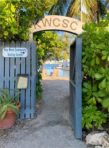 A wooden archway with 'KWCSC' welcomes visitors to the Key West Community Sailing Center, surrounded by lush green plants and a blue fence, overlooking water and dock chairs in the background.