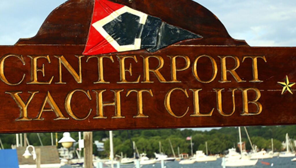 A wooden sign with gold lettering reads 'CENTERPORT YACHT CLUB' below a nautical flag, with sailboats and trees visible in the background.