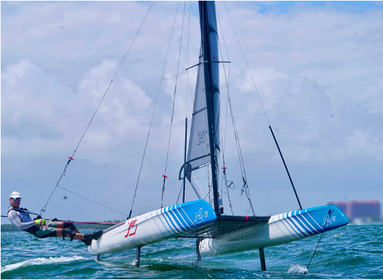 A person navigates a catamaran sailboat with blue and white hulls over the water on a partly cloudy day, leaning out over the edge while the boat's foils lift it above the waves.