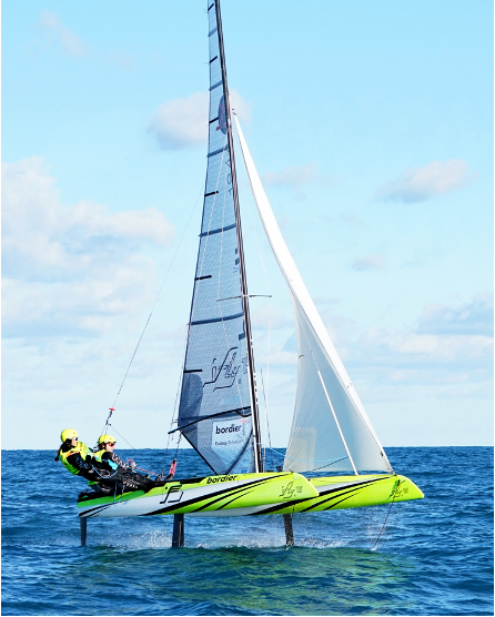 Two people in yellow helmets sail a bright green and white catamaran with lifted hulls, gliding above the blue water on hydrofoils under a partly cloudy sky.